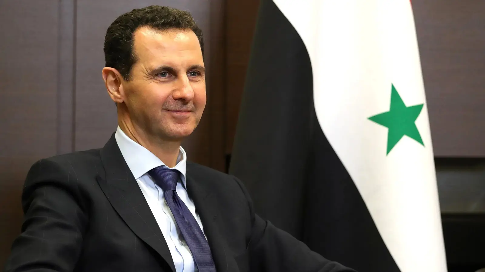 Bashar al-Assad seated beside a Syrian flag during a 2018 meeting in Russia