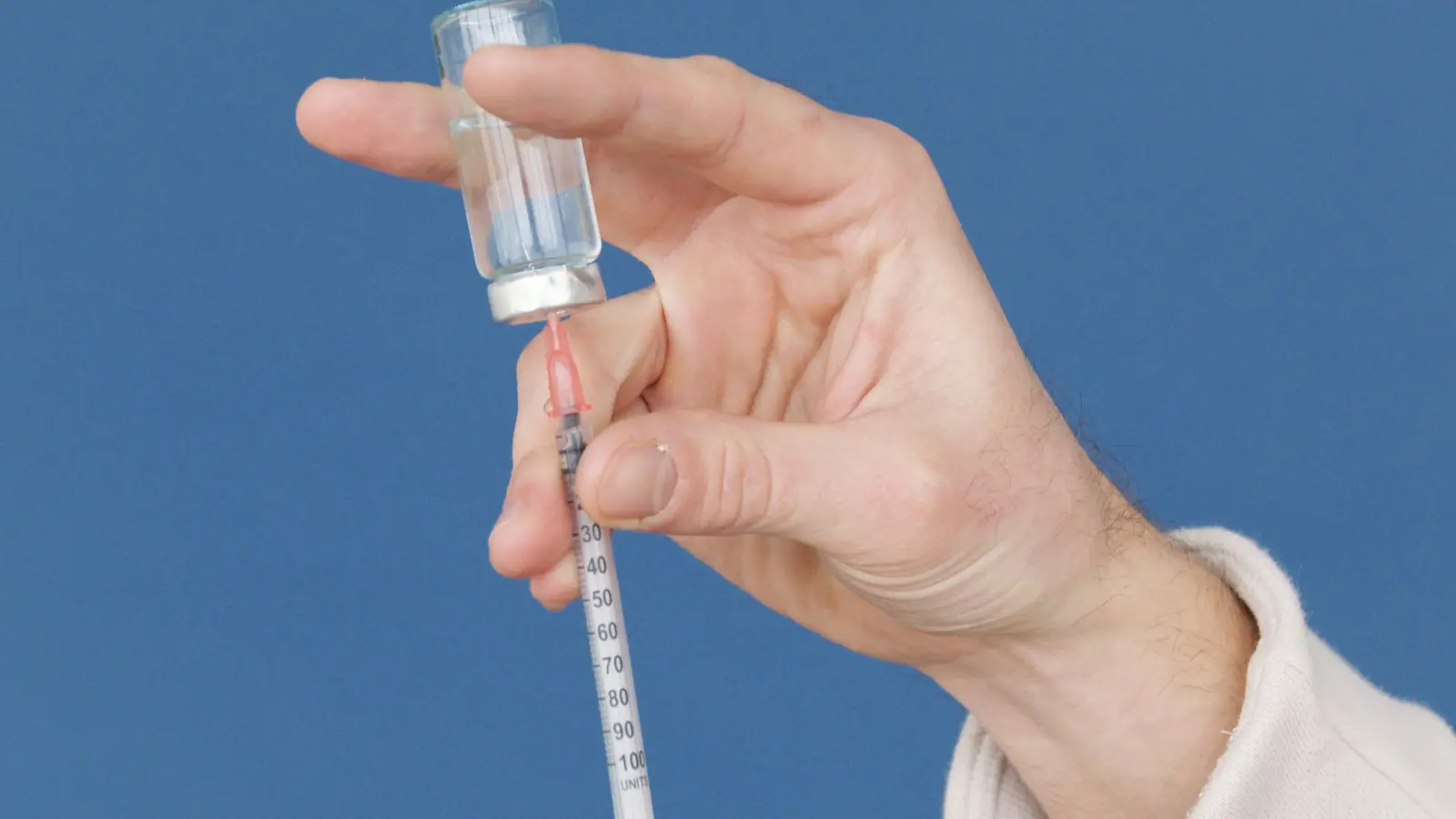 A medical worker draws vaccine from a vial into a syringe against a blue background